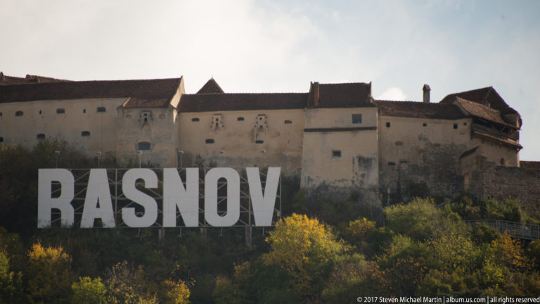 Large RASNOV hillside letters below the pale walls of Râșnov Fortress on an autumn-covered hill.