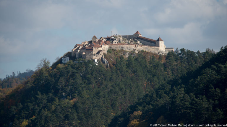 Râșnov Fortress clustered along the crest of a steep forested hill beneath a cloudy sky.
