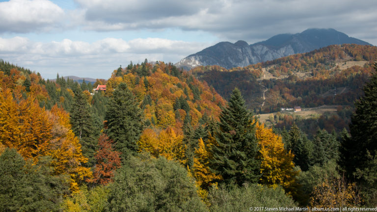 Autumn woodland, scattered buildings, and dark conifers below a rugged mountain ridge under heavy clouds near Bran.