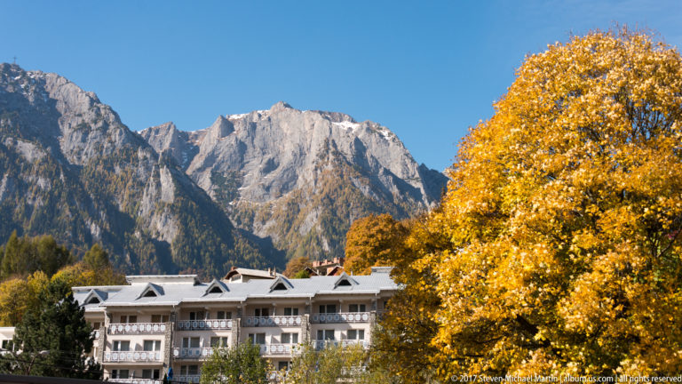 Multi-story buildings and golden autumn trees at the foot of a steep pale-gray Carpathian mountain massif.