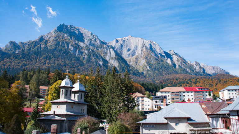 Small Orthodox church with twin pale domes among town buildings below a dramatic rocky Carpathian ridge.