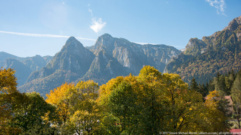 Jagged gray mountain peaks rise above golden autumn forest beneath a bright blue sky near Bran.