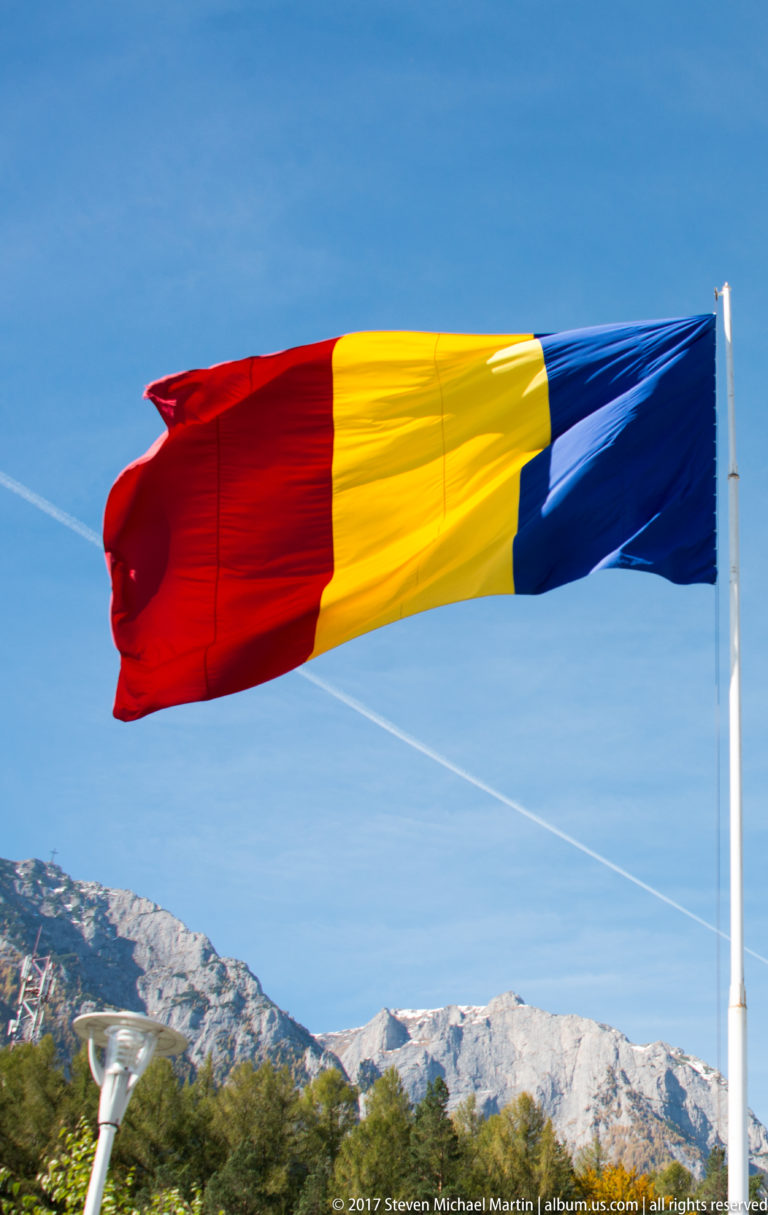 Romanian blue-yellow-red tricolor flying from a white pole with pale mountain ridges in the background.