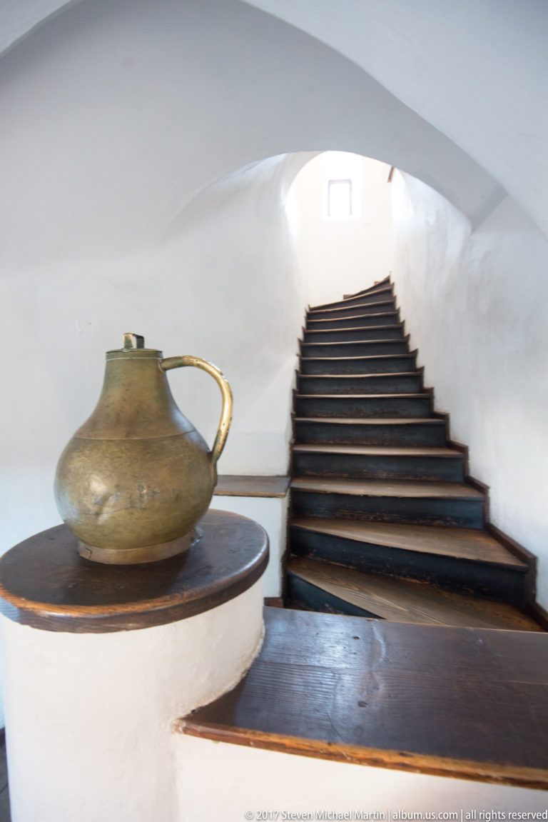 Large handled ceramic jug on a ledge beside a wooden staircase in a white-plastered interior.