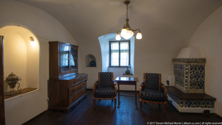 Furnished castle room with two upholstered chairs, wooden cabinet, small table, and blue-and-white tiled stove.
