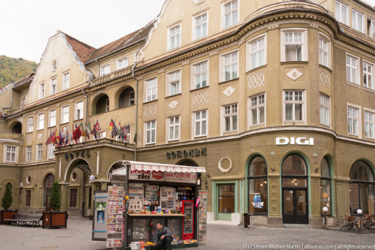 Curved historic corner building with tall windows, arcade openings, flags, storefronts, and a street kiosk.