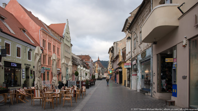 Narrow Brasov old-town street with cafes, pastel historic facades, shops, and distant pedestrians.