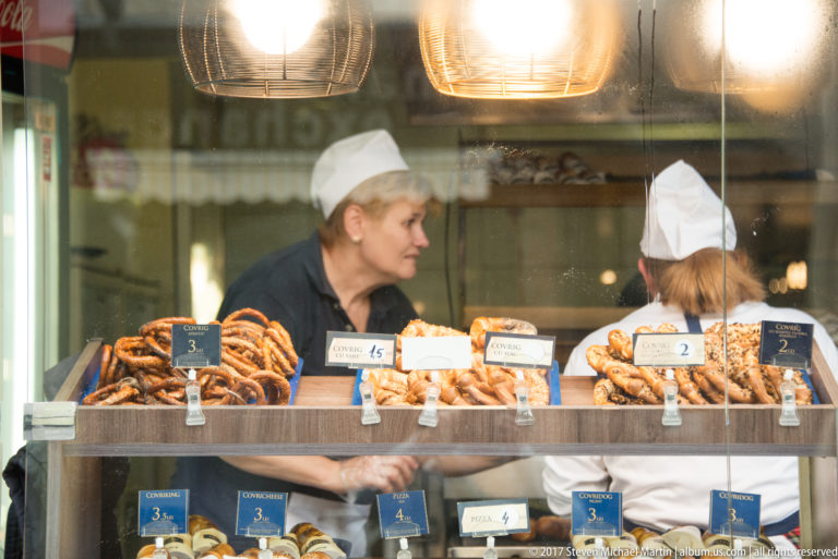 Two bakery workers behind a glass counter filled with pretzels and assorted pastries with price labels.