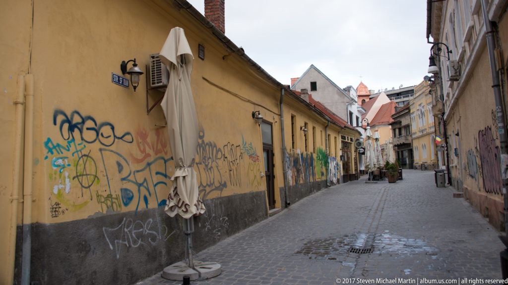 Narrow cobbled lane with graffiti-covered walls, closed umbrellas, lamps, and historic buildings in Brasov.