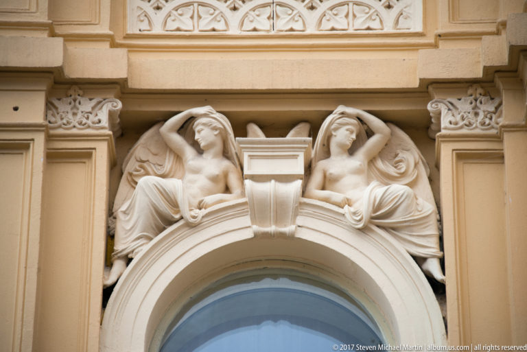 Two reclining female architectural figures with drapery positioned symmetrically above an arched window.