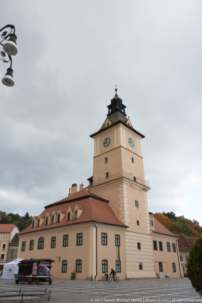 Brasov Council House with pale walls, red tiled roof, and tall clock tower under an overcast sky.