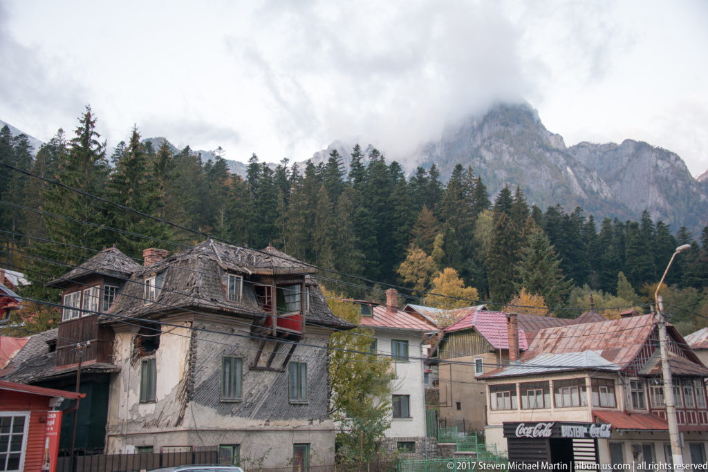 Weathered houses and balconies in a mountain settlement below forest and a clouded rocky ridge.
