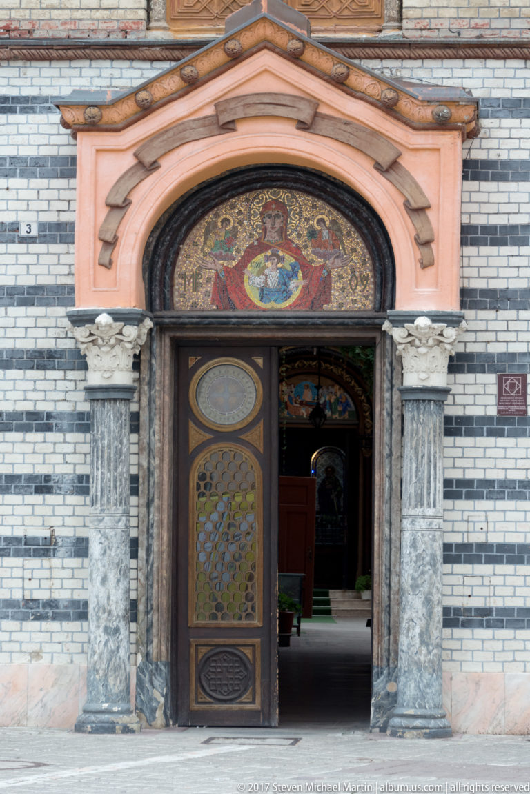 Open arched church doorway with stone columns, decorative doors, and a colorful mosaic above the entrance.