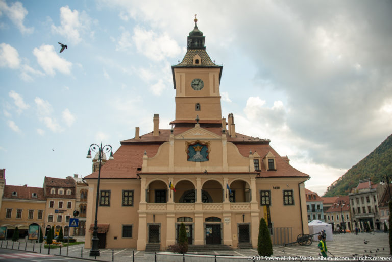 Frontal view of Brasov Council House with clock tower, upper arcade, heraldic emblem, and tiled roof.