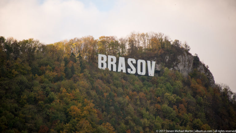 Large white BRASOV letters on a wooded autumn hillside with exposed rock.