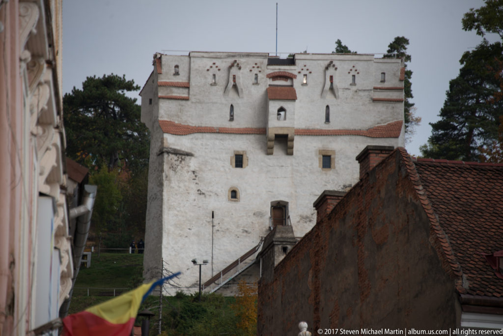 Whitewashed defensive tower with small openings rising above red tiled roofs on a wooded slope in Brasov.
