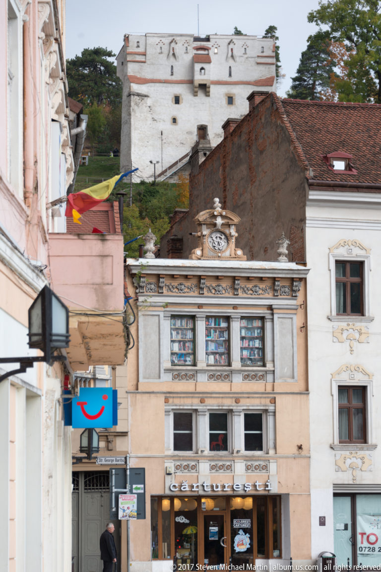 Decorated historic facade in Brașov with the whitewashed White Tower visible on the wooded hillside behind.