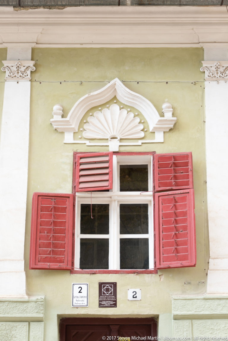 Historic window with red shutters and a white shell-shaped stucco pediment on a pale green facade.
