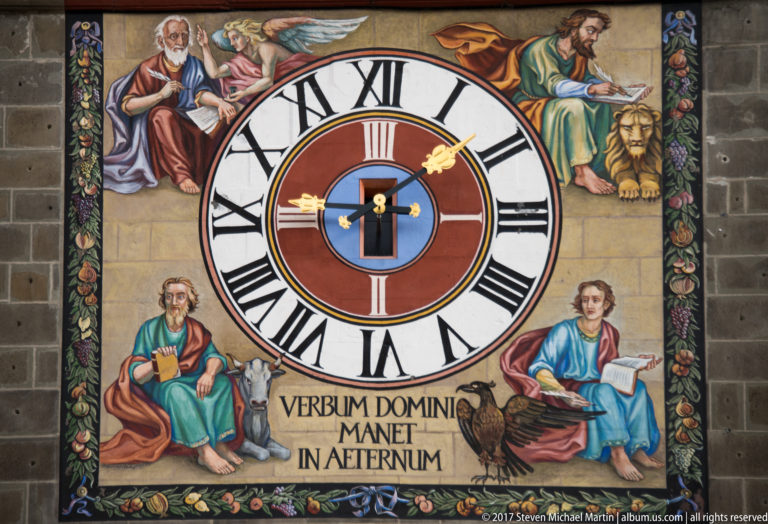 Painted clock with Roman numerals, religious figures, floral decoration, and the Latin inscription VERBUM DOMINI MANET IN AETERNUM.