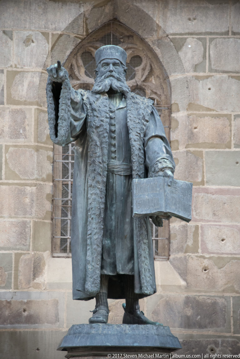 Bronze statue of Johannes Honterus holding an open book beside the stone wall of the Black Church in Brașov.