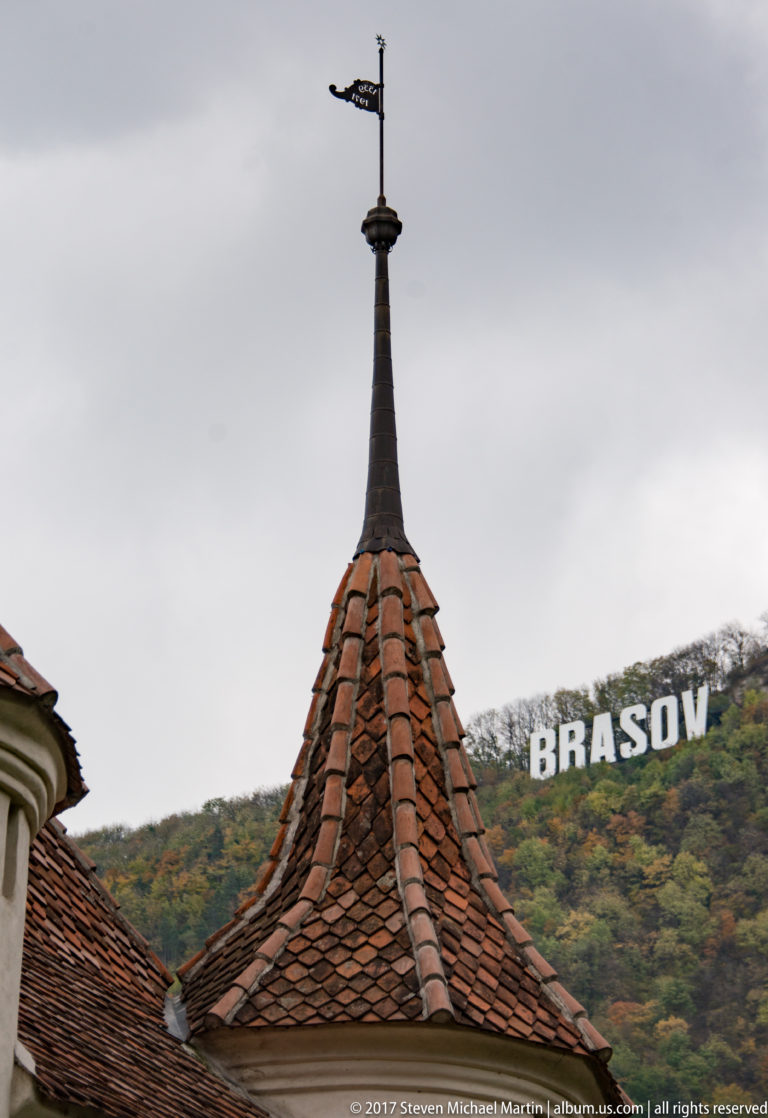 Red-tiled turret and weather vane in the foreground with the BRASOV hillside sign on wooded Tâmpa behind.