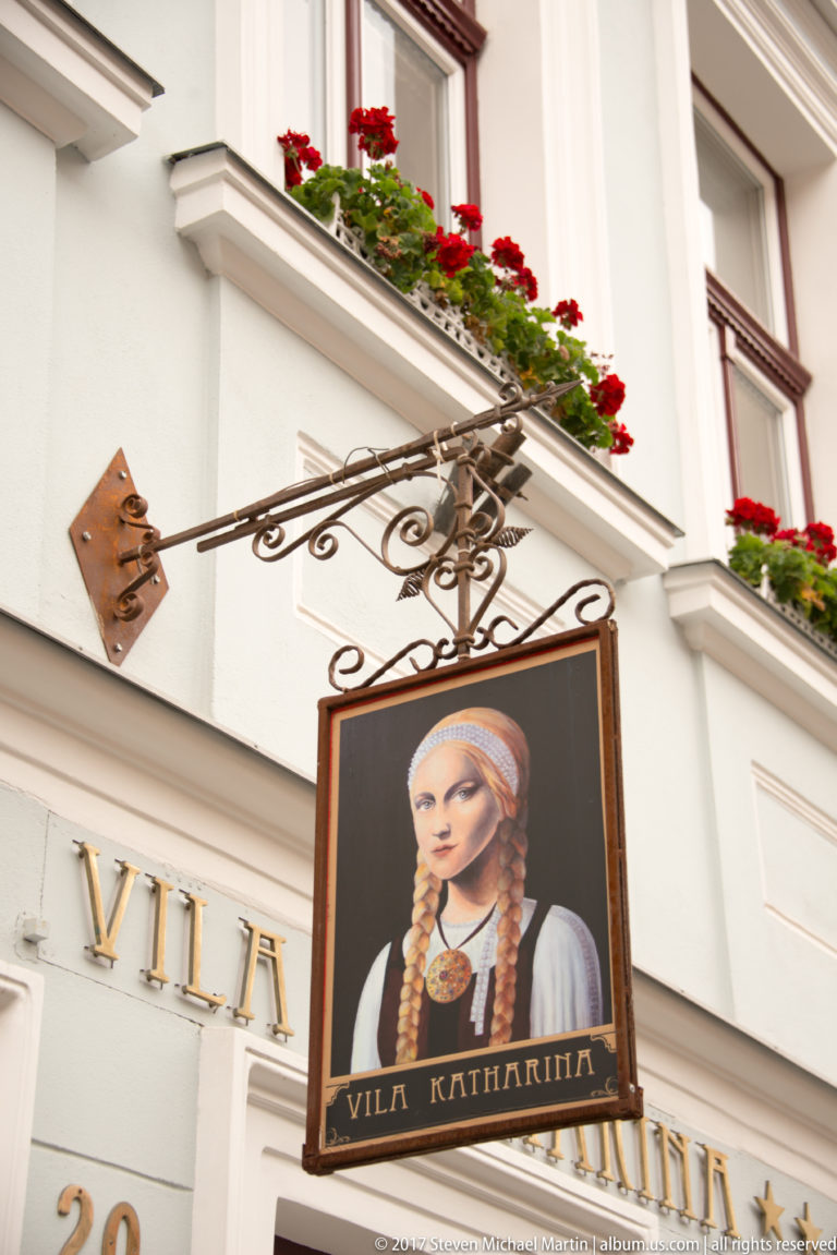 Hanging Vila Katharina sign with a portrait in historical dress beneath red geraniums on a white facade.