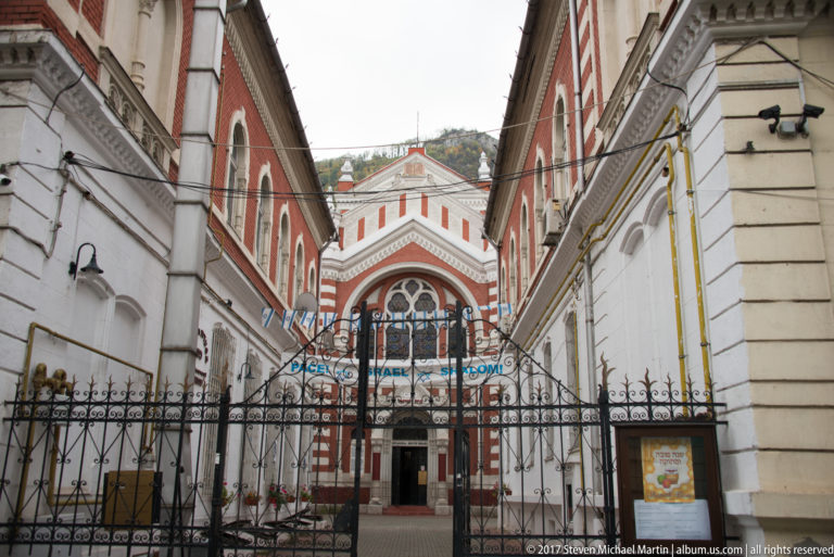 Beth Israel Synagogue seen through a gated courtyard, with red-and-white facade, arched windows, and blue-and-white decorations.