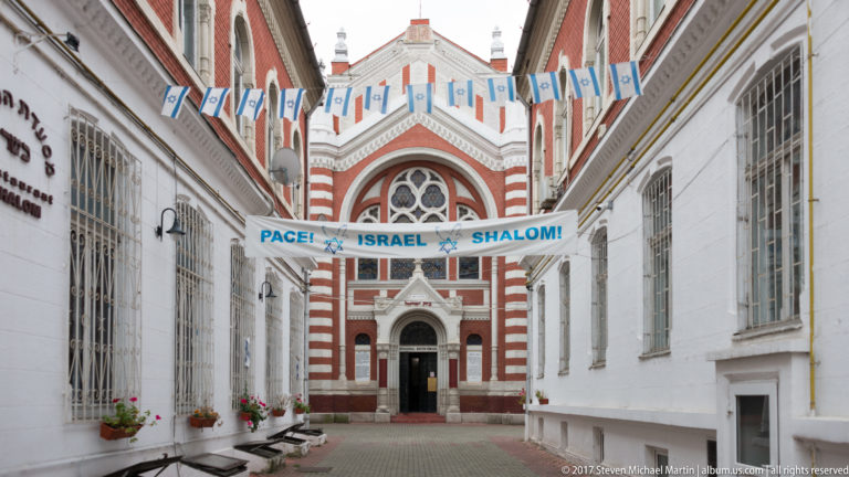 Narrow courtyard leading to the Beth Israel Synagogue beneath blue-and-white pennants and PACE, ISRAEL, SHALOM banners.