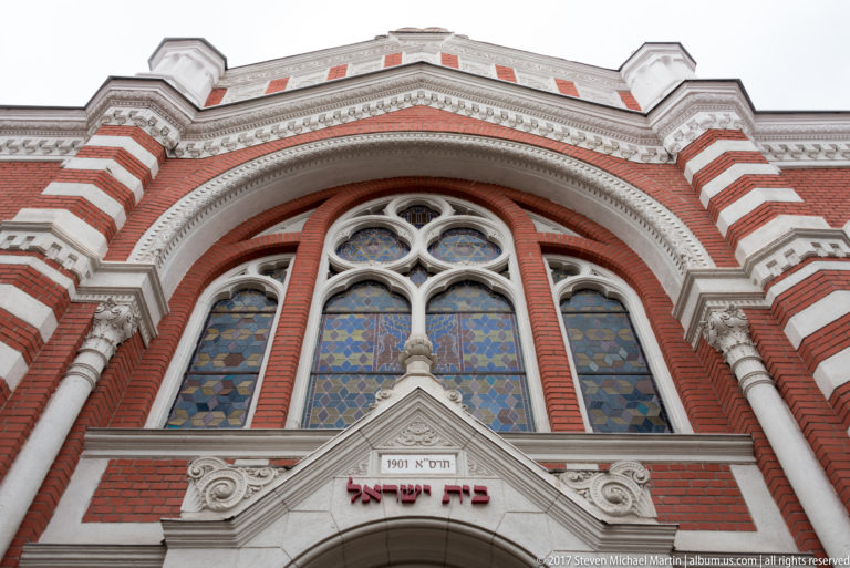 Upper facade of the Beth Israel Synagogue with red brick, striped masonry, stained glass, Hebrew lettering, and a 1901 date tablet.