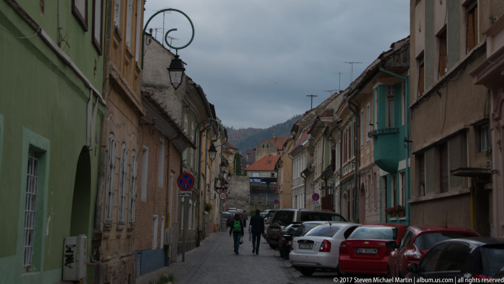 Narrow Brașov street with parked cars, pedestrians, historic houses, and wooded hills rising at the far end.