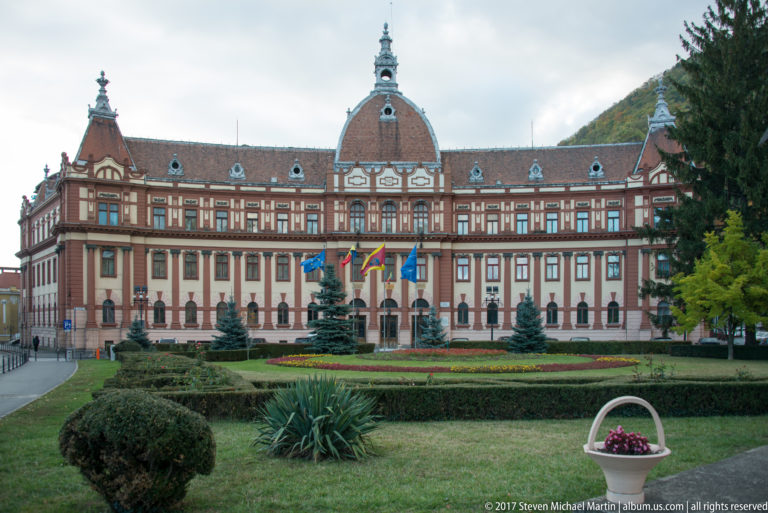 Symmetrical historic public building with tiled roof, central tower feature, flags, and formal gardens.