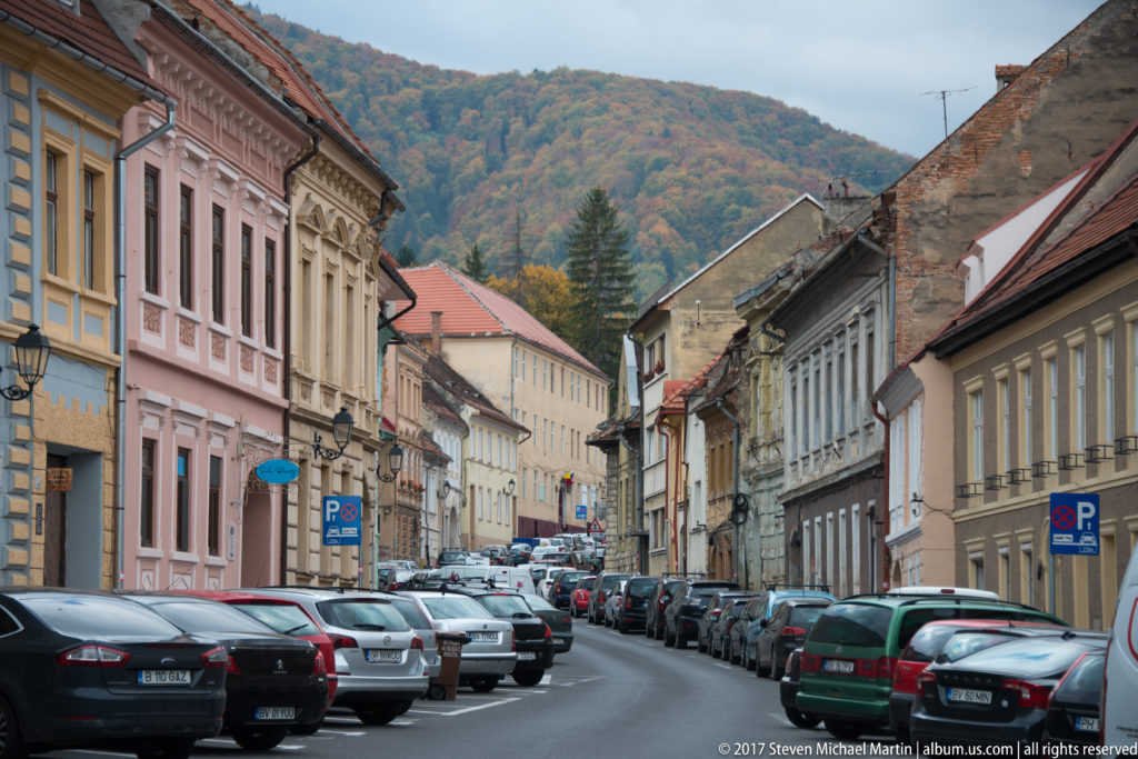 Brașov street lined with pastel historic buildings and parked cars, with an autumn wooded hill rising behind.