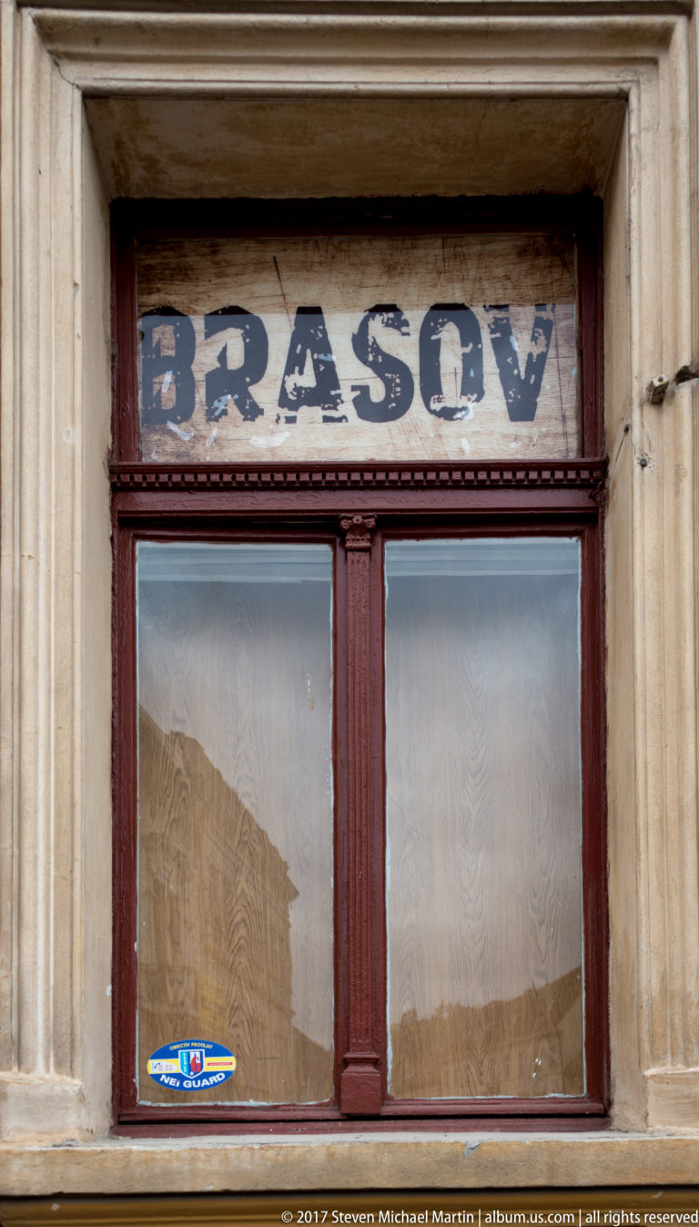 Faded BRASOV lettering above a dark red wooden shop window set within a pale stone surround.