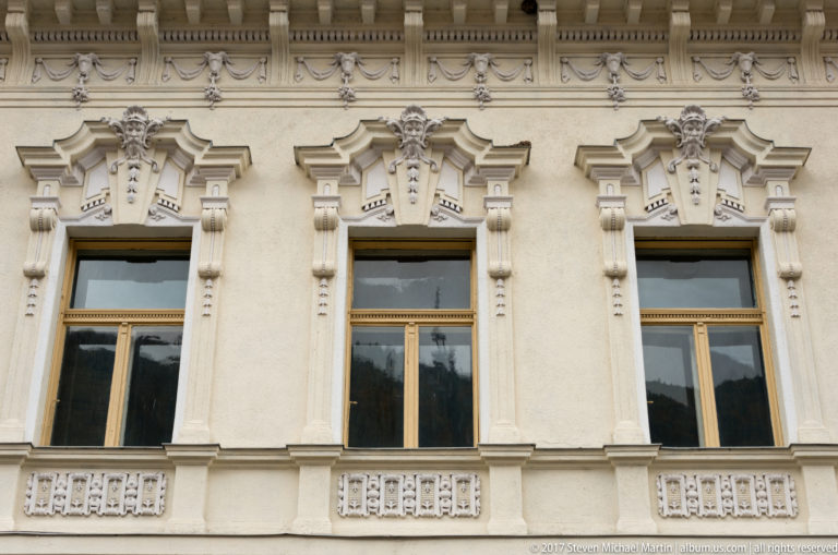 Three tall windows with elaborate cream stucco surrounds, sculpted pediments, and decorative panels on a historic facade.