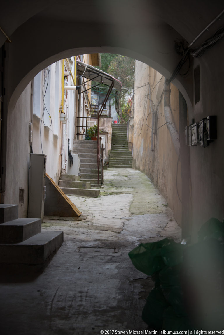 Shadowed archway opening to a worn courtyard with steps, exposed utilities, cracked plaster, and uneven paving.