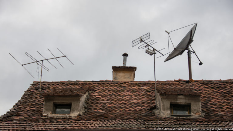 Red-tiled historic roof with dormer windows, brick chimney, multiple television antennas, and a satellite dish.