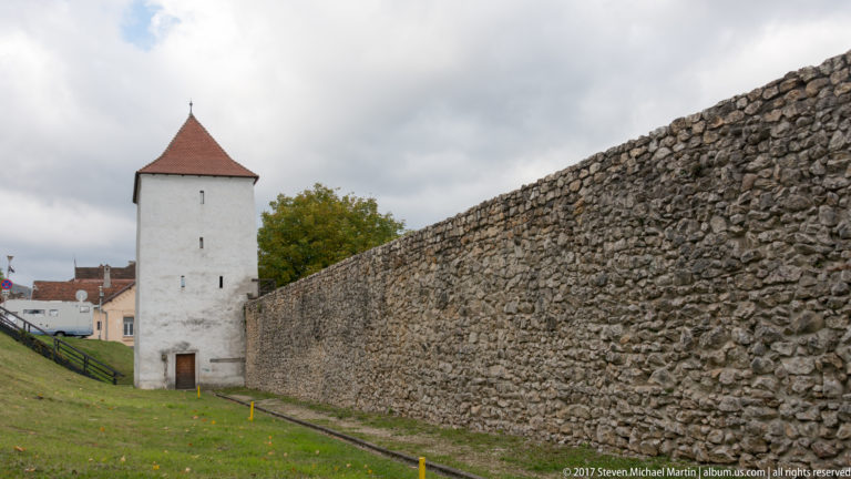 Long stone defensive wall leading to a square white tower with a red pyramidal roof beside a grassy strip.