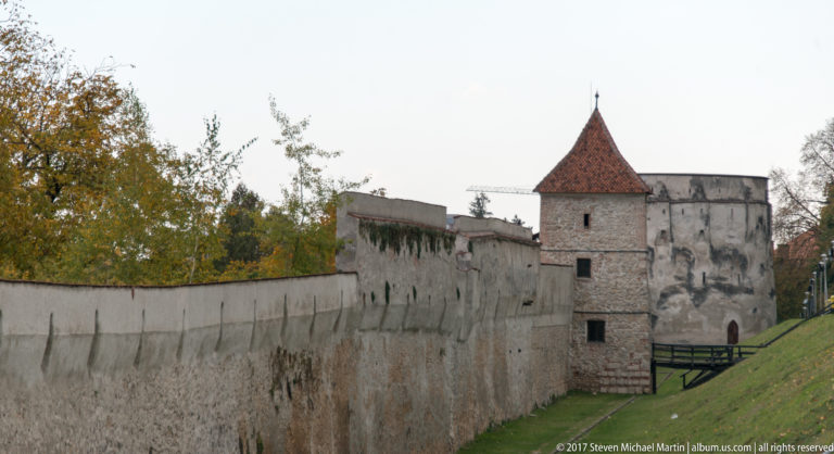 Stone city wall with a red-roofed defensive tower in front and a larger rounded fortification farther behind.