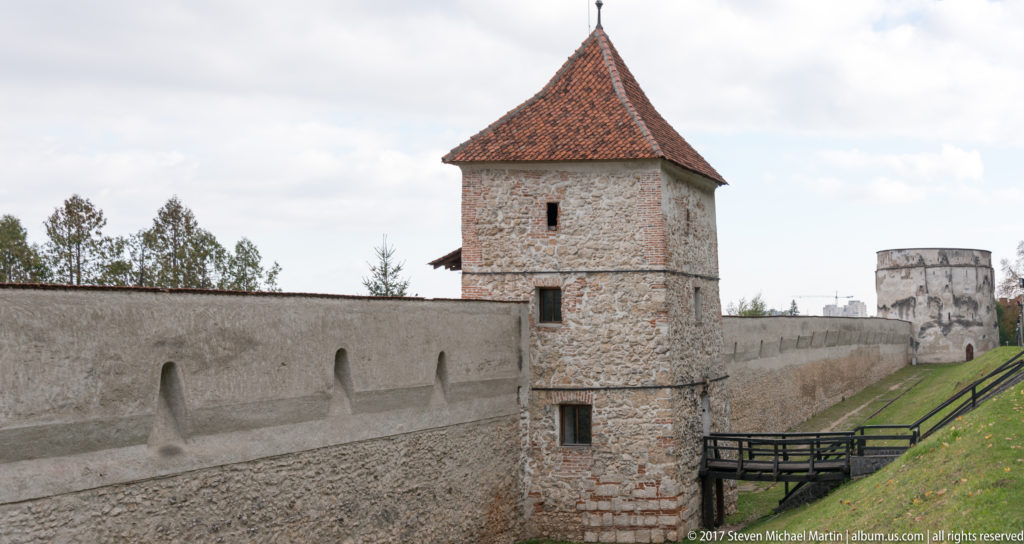 Square stone defensive tower with a steep red roof built into Brașov’s city wall, with a rounded bastion in the distance.