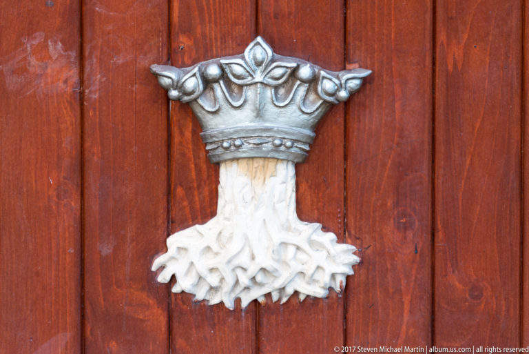 Crowned white tree with exposed roots mounted as a heraldic emblem on dark red wood.