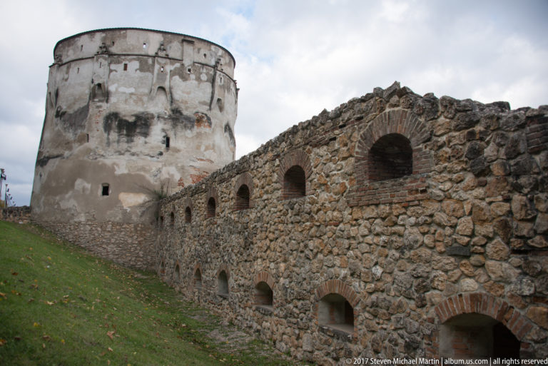 Rough stone fortification wall with rows of arched openings leading toward a large rounded bastion.