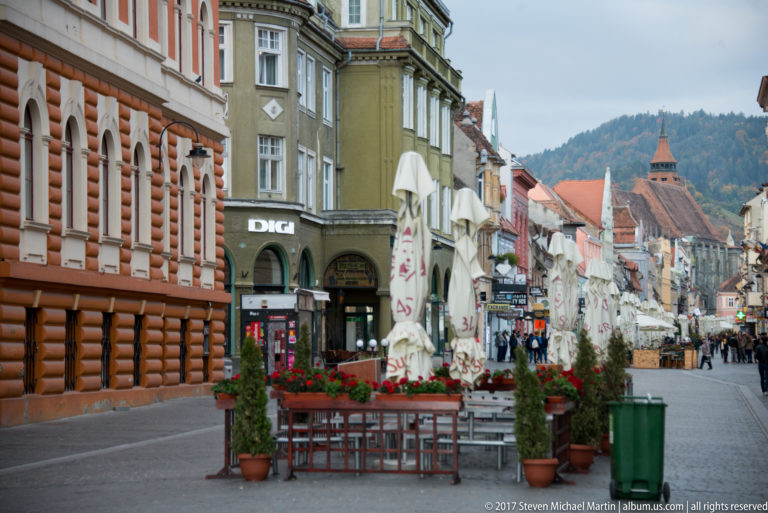Pedestrian street with cafes, planters, historic facades, and a distant church roof in Brasov.