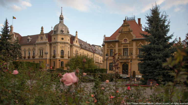 Ornate historic buildings with a domed corner tower and steep red roof behind roses and evergreens.