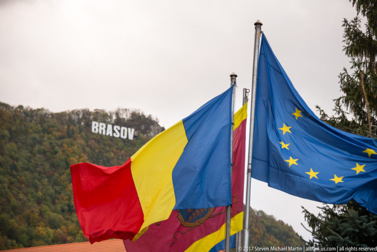 Romanian and European Union flags in front of the wooded hill carrying the BRASOV sign.