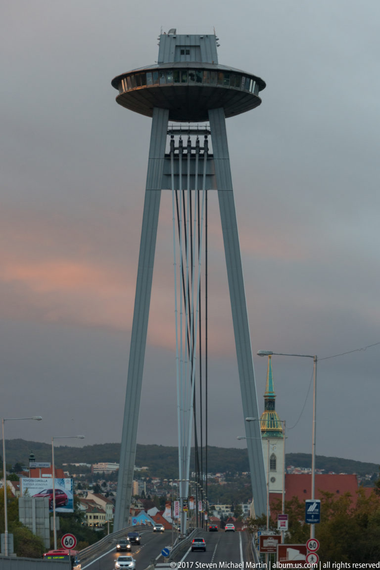Most SNP bridge pylon and saucer-shaped observation deck rising above traffic with Bratislava buildings in the distance.