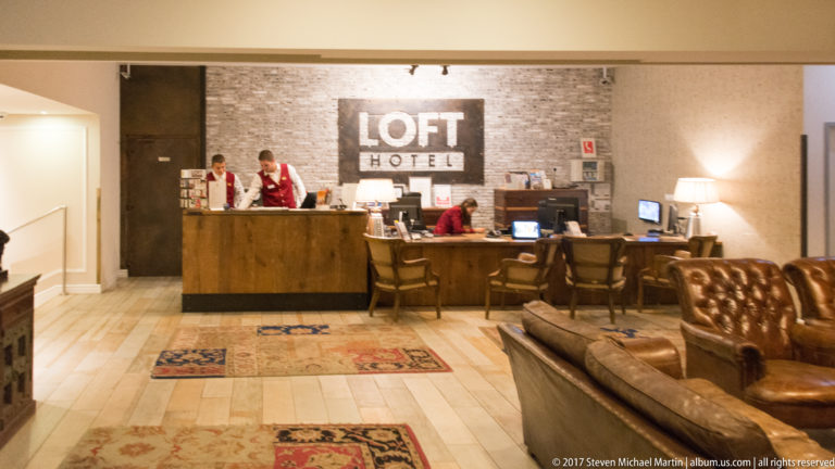 Loft Hotel reception desk with staff, large wall sign, warm lamps, rugs, and brown leather lounge seating.