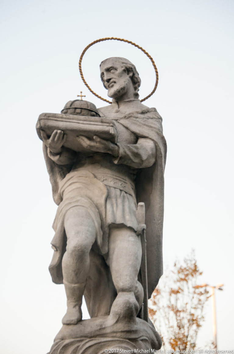 Stone statue of a haloed male saint holding a broad covered object against the sky.