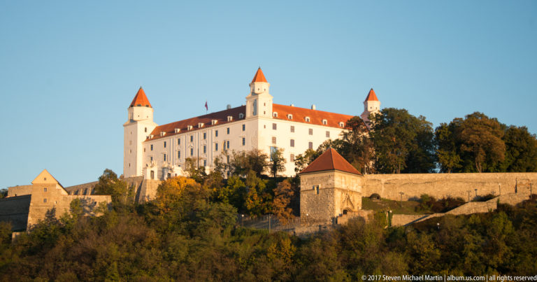 Bratislava Castle on its fortified hill, with four corner towers, red roofs, walls, and wooded slopes.