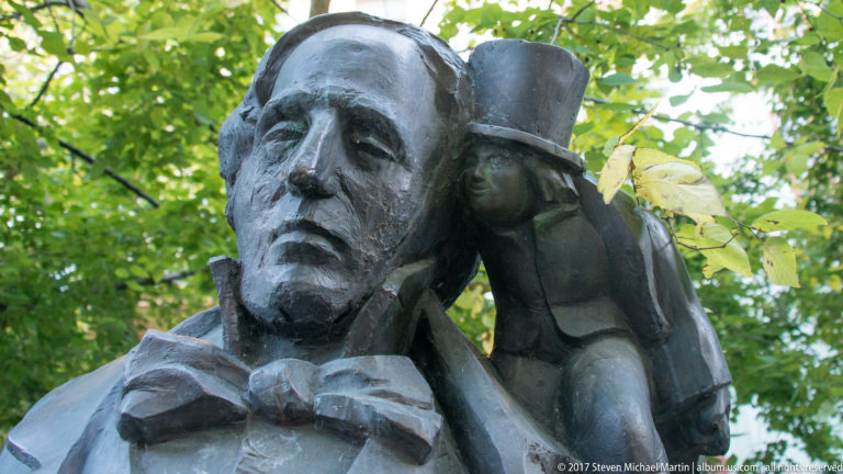 Bronze sculpture of a solemn man with a much smaller top-hatted figure leaning beside his head.