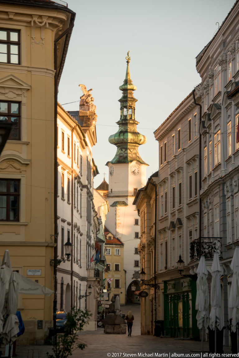 Narrow Bratislava Old Town street leading toward a pale tower, with ornate roof sculpture and historic façades.